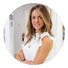 Woman with long, light brown hair wearing a white sleeveless top stands with arms crossed and smiles at the camera in a brightly lit indoor setting, radiating confidence in her oral health and happiness.