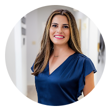 A woman with long brown hair, wearing a dark blue blouse, stands smiling in a brightly lit hallway, radiating the warmth often found in family dentistry.