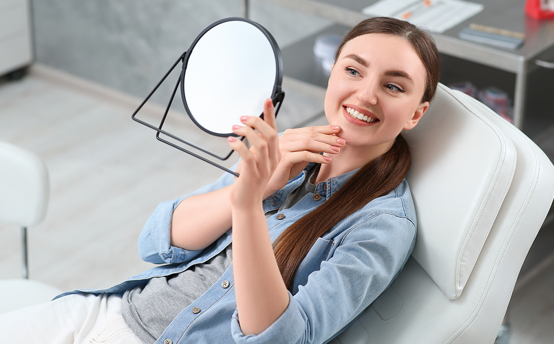 A woman sits in a dental chair, smiling and looking at her reflection in a handheld mirror after her dentist visit.